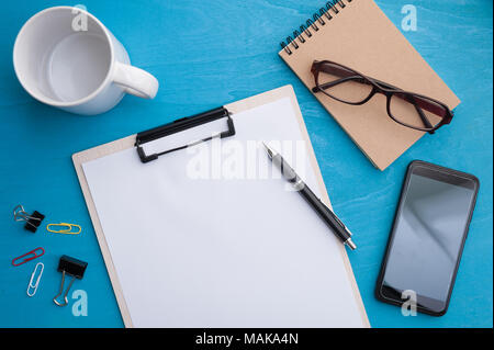 A4 size clipboard, smartphone, small notebook and coffee cup on blue wood table in workplace. background with blank area for text or message. Stock Photo