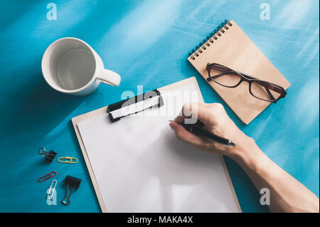 Young Asian male hand writing on white paper on a4 size clipboard on blue wood table in workplace. freelance worker lifestyle concept Stock Photo
