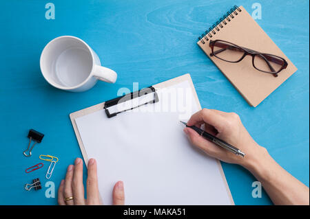 Young Asian male hand writing on white paper on a4 size clipboard on blue wood table in workplace. freelance worker lifestyle concept Stock Photo