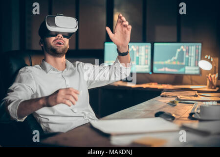 Young bearded man trader at office sitting at table looking at universe in virtual reality headset excited Stock Photo