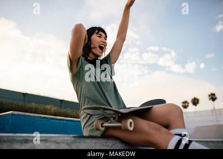 Smiling urban girl sitting on skateboard ramp in skate park having fun. Female skateboarder enjoying herself outdoors at skate park. Stock Photo