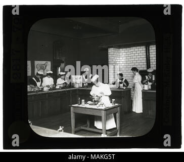 Horizontal, black and white photo of young women, in aprons and ruffled caps, standing at cooking stations. Title: Educational Museum Lantern Slides: Cooking- McKinley High School.  . circa 1904. Stock Photo