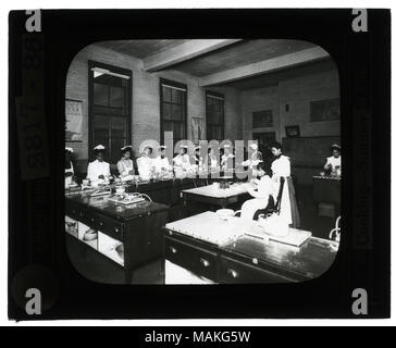 Horizontal, black and white photo of young African-American women, in aprons and ruffled caps, standing at cooking stations. Title: Educational Museum Lantern Slides: Cooking- Sumner High School.  . circa 1904. Stock Photo