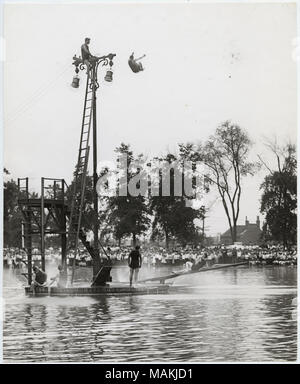 Vertical, black and white photograph showing male divers jumping from a high-dive platform that has been improvised at the top of a light pole at Fairground Park Pool. One diver is shown in mid-air, while another is perched atop the light pole. A ladder leads from the diving board platform to the top of the light pole. The diving board platform is in the middle of the pool and also features two low diving boards and a lower high-dive diving board. Four other swimmers are watching the diver from the platform, and a large crowd watches from the shore in the background. Title: Divers improvise a  Stock Photo