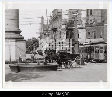 Horizontal, black and white photograph showing horses drinking from the trough at the base of the Grand Avenue water tower. The horse drinking from the trough is pulling a wagon. The base of the water tower is on the left. Several streetcars can be seen on the right. Two and three-story brick commercial buildings, pedestrians, and a car can be seen in the background. Title: Horse drinking at the trough at the base of the Grand Avenue water tower.  . circa 1910. Holt, Charles Clement, 1866-1925 Stock Photo