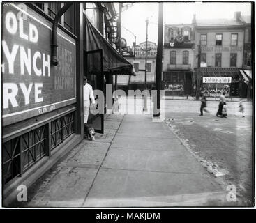 Horizontal, black and white photograph showing a man letting a dog out the back door of a saloon at 1151 South Broadway. The man is dressed in white, possibly wearing a white coat or apron. A sign painted on the side of the saloon advertises Old Lynch Rye. Several blurry figures, including a woman and children, can be seen crossing the street on the right side of the photo, probably Hickory Street. Signs for a milliner and shoe store can be seen across South Broadway. Title: Man letting a dog out the back door of a saloon at 1151 South Broadway.  . circa 1907. Stock Photo