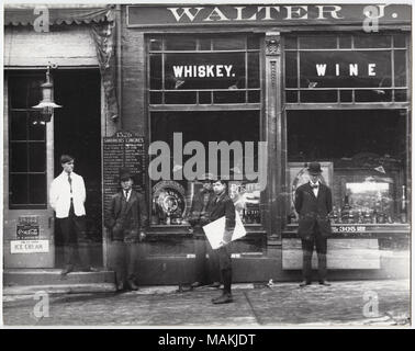 Horizontal, black and white photograph showing Walter J. Noble Whisky and Wine at 1328 Market Street and Mielke's Lunch Stand at 1326 Market Street. A man wearing a white coat stands in the doorway of Mielke's Lunch Stand, and a menu listing sandwiches and lunches is to the right of the doorway. Three other men and a newsboy are standing in front of the Walter J. Noble Whisky and Wine display windows. The windows show a variety of bottles and advertisements, including ads for Pabst and Old Crow Distillery. This image is a detail of image number 34877. Title: Men standing in front of Walter J.  Stock Photo