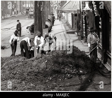 Horizontal, black and white photograph showing a street crew backfilling after laying new underground utilities on Sarah Avenue near Washington Boulevard. A mound of dirt sits in the foreground, covering the sidewalk. Behind the pile, five workers are shoveling dirt. Additional sections of pipe are laid out along the curb. Wagons and pedestrians can be seen in the background. Shops along the left side of the image advertise coffee and plumbing services. A handwritten note on the back of the print, possibly from the School Sisters of Notre Dame, reads: 'Washington and Sarah / about 1914-1915.' 