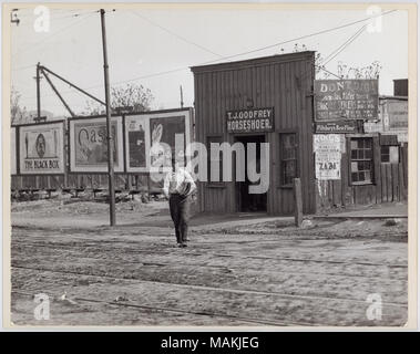 Horizontal, black and white photograph showing a boy or young man walking away from T. J. Godfrey Horseshoer, located at 3968 Chouteau. The boy is carrying what appears to be a stack of newspapers, and he is about to cross two sets of streetcar tracks that run through the foreground. T. J. Godrey's is a one story wooden building, with advertisements plastered along the side. Several large billboards are to the right of the building. Advertisements promote the Black Box, Oasis Cigarettes, Jag. Alder's plumbing services, Pillsbury's Best Flour, and King's Theatre. Title: T. J. Godfrey Horseshoer Stock Photo