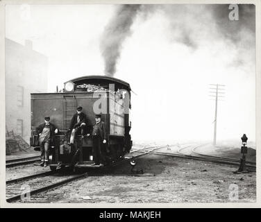 Horizontal, black and white photograph showing three men on the back of a steam switch engine in a rail yard. Two of the men stand on a running board, while the third is perched on a rail or pipe. One man may be the conductor, as he wears a uniform hat and a suit, while the other two men wear workmen's clothing. A headlight is at the top of a short ladder, and coal piled in the bunker can be seen behind it. A plume of smoke billows away from the train. Several railroad tracks criss-cross in the background. A handwritten note on the back of the print, probably from the School Sisters of Notre D Stock Photo