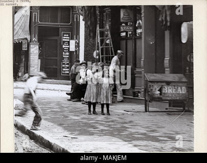 Horizontal, black and white photograph showing two young girls standing on the sidewalk near the corner of Broadway and Cerre. A small group of men is seated behind them, and a boy is running onto the sidewalk at the left edge of the photo. A liquor store or saloon in the background advertises Old Governor Rye whiskey, Old Capitol bourbon, Stifel beer, and other liquors. A large box with a bread advertisement is on the sidewalk to the right of the girls. Photograph is a detail of image number 25971. Title: Two girls standing on the sidewalk in front of a small group of seated men near the corn Stock Photo