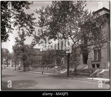Horizontal, black and white photograph showing Washington near Grand. The street runs along the lower edge of the photograph, and a sidewalk can be seen on the far side of the street. A low stone wall borders much of the sidewalk. A house with ornamental carvings around the windows and doors is on the right side of the image, and a man can be seen passing in front of it on the sidewalk. Other buildings are only partly visible behind trees and foliage. A handwritten note on the back of the print, possibly from the School Sisters of Notre Dame, reads: 'Washington near Grand / Print by Dr. Sherwi Stock Photo