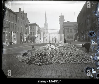 Horizontal, black and white photograph showing a large pile of broken stone on a cobblestone street. There is one man at work on the right side of the picture, and another man seated and looking at the viewer from the back of the rubble pile. In the distance there are pedestrians and wagon traffic and a church with a steeple visible. Title: Street workers beside a pile of rubble on a cobblestone street.  . circa 1910. Holt, Charles Clement, 1866-1925 Stock Photo