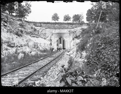 Horizontal, black and white photograph showing two men with cameras positioned on train tracks next to a tunnel. One photographer has the camera over his shoulder and is facing the viewer. The other photographer has his back to the viewer and is in the process of taking a picture of the tunnel. An adult and child are standing near the tunnel's entrance. Rocks and foliage can be seen on either side of the tracks and above the tunnel. Title: Photographers on the tracks at the Meramec Highlands Tunnel.  . 1895. Stock Photo