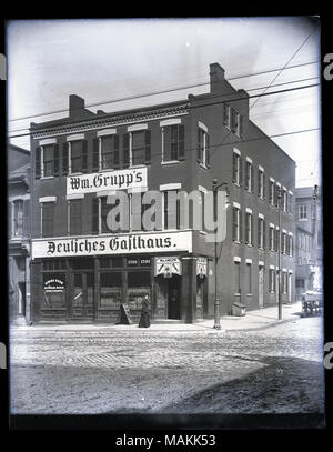 Vertical, black and white photograph showing William Grupp's saloon and boarding house, Grupp Hall, located at 1701 Market Street at the corner of Market Street and 7th Street. The large sign on the three-story brick building is in German and reads: 'Wm. Grupp's Deutsches Gasthaus.' A sign in the first floor windows reads: 'Wm. Grupp's Saloon / Union Bar.' Other signs advertise rooms, meals, and Excelsior beer. A woman is standing near the corner entrance. A cobblestone street runs through the foreground. Title: William Grupp's saloon and boarding house, Grupp Hall, 1701 Market Street.  . betw Stock Photo