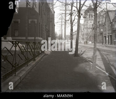 Horizontal, black and white photograph showing a view looking down the brick sidewalk of an unidentified street. Both sides of the street have residential buildings next to large, stone buildings, possibly churches. In the foreground on the left, there is a wrought-iron fence enclosing a small front yard. The street is on the right. Title: View looking down an unidentified street.  . between 1900 and 1920. Stock Photo