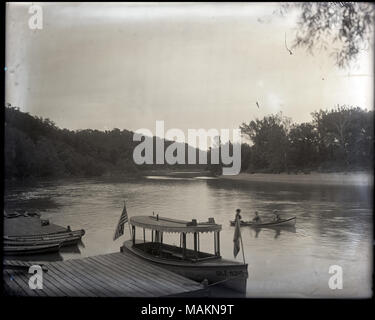 Horizontal, black and white photograph showing a boat called 'Idle Hour' tied up at a dock in the foreground. Behind 'Idle Hour', two men and a woman are boating in a small row boat. Trees line the riverbanks, and the river curves to the right in the background. Title: Recreational boats on a river.  . between 1910 and 1920. Stock Photo