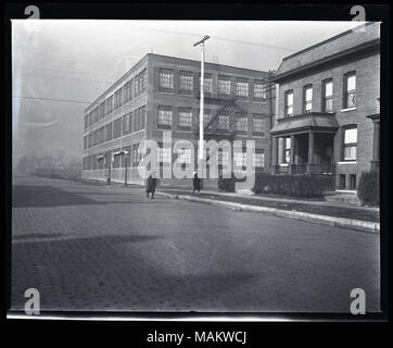 Horizontal, black and white photograph showing a three-story factory or warehouse across the street from a two-story multi-family residential building. The factory has large windows and a fire-escape on the side of the building. The residence is a two-story brick building, probably an apartment building, and has a small porch. A small poster with a light background and a cross in the center is displayed in one of the windows, possibly a Red Cross Service Flag given out during World War I. A cobblestone street runs through the foreground and two pedestrians can be seen. Original envelope was la