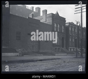 Horizontal, black and white photograph showing a row of two-story brick residential buildings next door to an unidentified industrial or commercial building. The residential buildings have decorative brickwork along the roofline and around the windows. It is unclear if the buildings are single family homes or multi-family apartment buildings. A small poster with a light background and a cross in the center is displayed in one of the windows, possibly a Red Cross Service Flag given out during World War I. The industrial or commercial building is also brick. There is a set of large double doors  Stock Photo