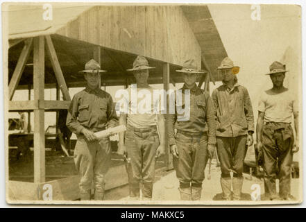 Horizontal, sepia photograph showing five uniformed men posing for a group picture standing in front of a wooden structure with open sides. The men are holding various tools, including hammers and an object that is possibly a carpenter's plane. A caption on the back reads 'Co. I Carpenters. / Lt. A. Skinker in Charge.' Title: 'Co. I Carpenters.'  . between circa 1916 and circa 1918. Stock Photo