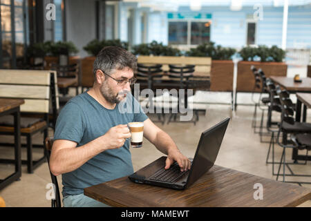 Freelance and working online concept. Man working with a black laptop, while drinking latte in a coffee shop. Stock Photo