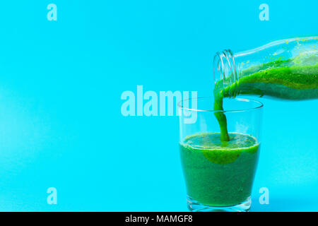 Process of Pouring from Bottle to Glass of Green Fresh Smoothie from Leafy Greens Vegetables Fruits. Apples Bananas Kiwi Zucchini Spinach on Light Blu Stock Photo