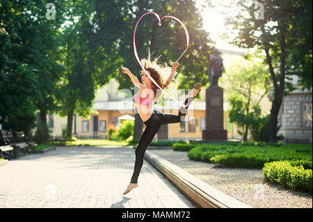Ivano-Frankivsk, Ukraine - 1 June 2015 : Young pretty woman with stunning figure is dancing with red ribbon in morning city park. Woman making pirouettes on garden bed near green plantings. Stock Photo