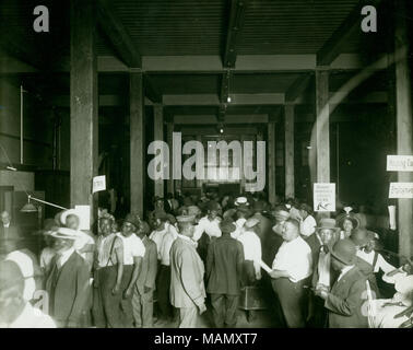 Horizontal, black and white photograph showing a large open room where a group of African Americans, mostly men, There is a large central aisle in the room. Along the left side men are standing in a line, in the center people are exiting the room, and along the right side people are seated or waiting. There are some signs posted on poles in the room reading 'Report Property Losses Here', 'Housing Con[ditions]', and 'Employment.' The photograph was possibly taken at the Municipal Lodging House (110 South 12th Street). Title: Group of African American men reporting property losses around time of