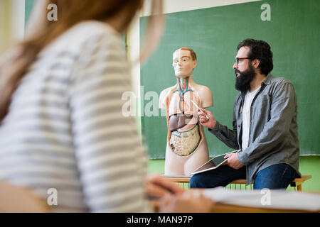 Young male hispanic teacher in biology class, holding digital tablet and teaching human body anatomy, using artificial body model. Stock Photo