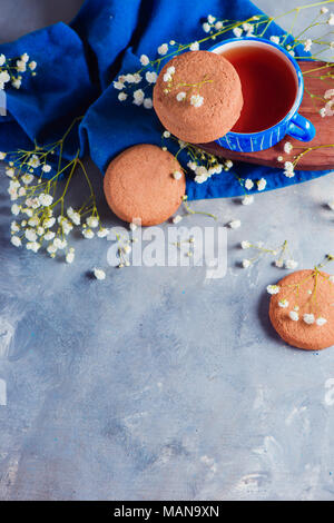 Morning scene with a small blue teacup and oatmeal cookies on a blue linen napkin. High key snack concept with homemade pastry and spring gypsophila f Stock Photo