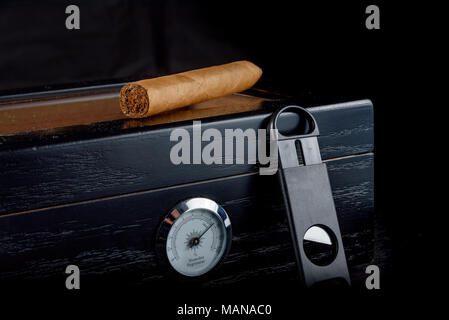 A rich smelling cigar on a black humidor next to a straight cigar cutter. Black background Stock Photo