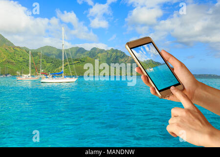 Woman hand hold and touch screen smart phone over Beautiful sea and sailing boat in Moorae Island at Tahiti , PAPEETE, FRENCH POLYNESIA in the morning Stock Photo