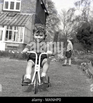 1960s, historical picture of a small boy sitting on his metal tricycle outside in a garden, England, UK. Stock Photo