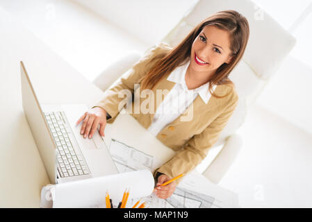 Smiling young woman architect analyzing blueprint on laptop at desk in office.Top view. Stock Photo