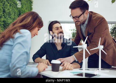 Smart qualified skilled young engineers looking at the project of alternative energy with miniatures of windmill turbines at the table. Stock Photo