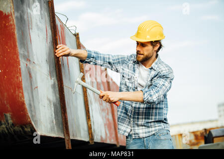 strong man with beard in plaid shirt, very concentrated, hammering or trying to repair some rusty objects that require attention, outside on a site Stock Photo