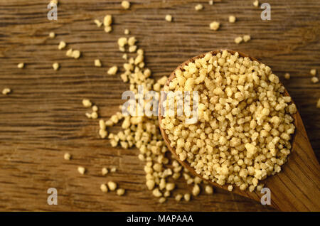 Overhead close up of couscous grains, filling wooden spoon and spilling on to oak surface below. Stock Photo