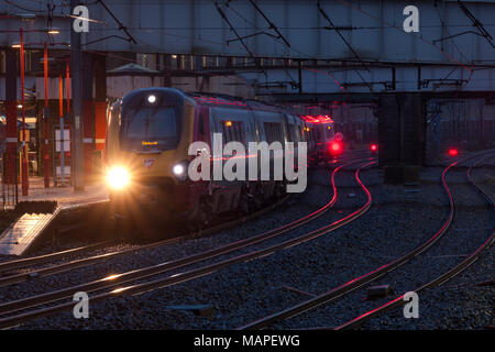 A Virgin Trains west coast Voyager train at Lancaster railway station on the West Coast main line  with red signals glinting off the track at night Stock Photo