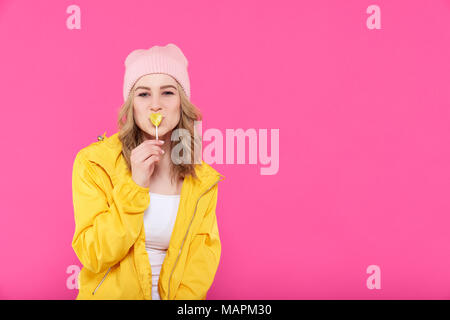 Beautiful trendy girl in colorful clothes and pink beanie kissing heart shaped popsicle. Attractive cool young woman fashion portrait. Stock Photo