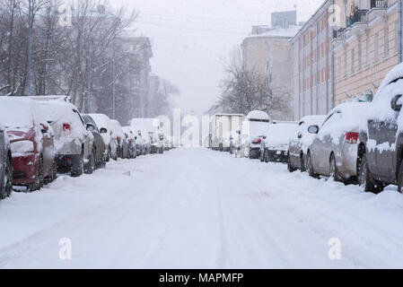 Snowfall in empty street. Rows of parked cars covered with snow. Buildings and park can be seen through spots of falling snowflakes. Winter in city Stock Photo