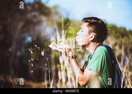 Young man blows away seeds from his hands while a hike in the forest. A hiker who wears backpack walks among tall dry grass in the sunny afternoon. Stock Photo