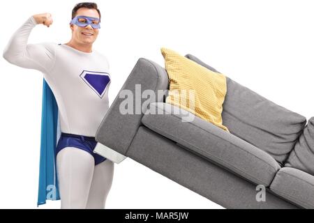 Young man in a superhero costume lifting a sofa and flexing his biceps isolated on white background Stock Photo
