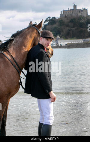 Handsome Male Horse Rider standing walking his horse, wearing flat cap, white trousers and black boots on beach in front of St Michael's Mount Stock Photo