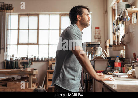 Skilled young woodworker leaning on a workbench deep in thought Stock Photo