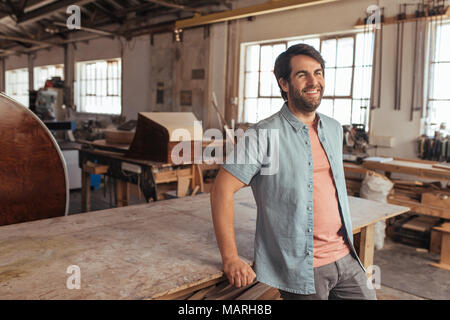 Smiling young woodworker leaning on a table in his workshop Stock Photo