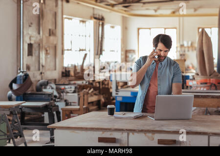 Woodworker using a phone and laptop in his large workshop Stock Photo