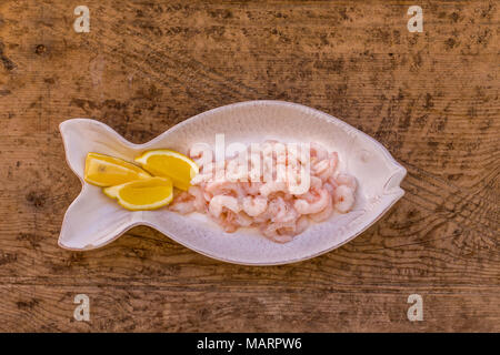 Upper view shot of raw shrimps and lemon in fish shaped rustic seramic plate on wooden tabletop. Stock Photo