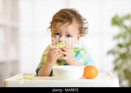 Cute baby 1 years old sitting on high children chair and eating fruits alone in white kitchen Stock Photo