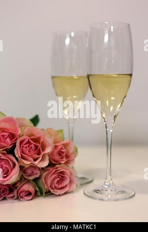 Two champagne flutes / sparkling wine glasses photographed on a white table with a bouquet made of light pink roses. Luxurious celebration! Stock Photo