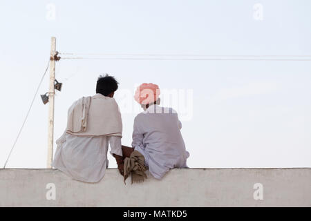 Two men dressed in white clothing, one wearing a pink turban, sitting on a wall at the stadium at the Pushkar Camel Fair, Pushkar, Rajasthan, India. Stock Photo